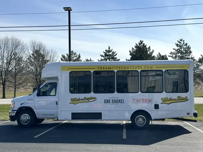 A white shuttle bus wrapped with the Who's on Layton, Oak Barrel, Tavern and Who's on Third logos, parked beside a lot on a sunny day