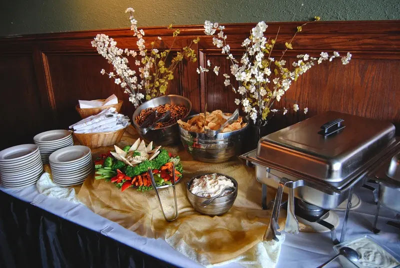 A buffet table with chafing dishes, pretzels and a veggie platter beside vases of spring blossoms in a wood-paneled room