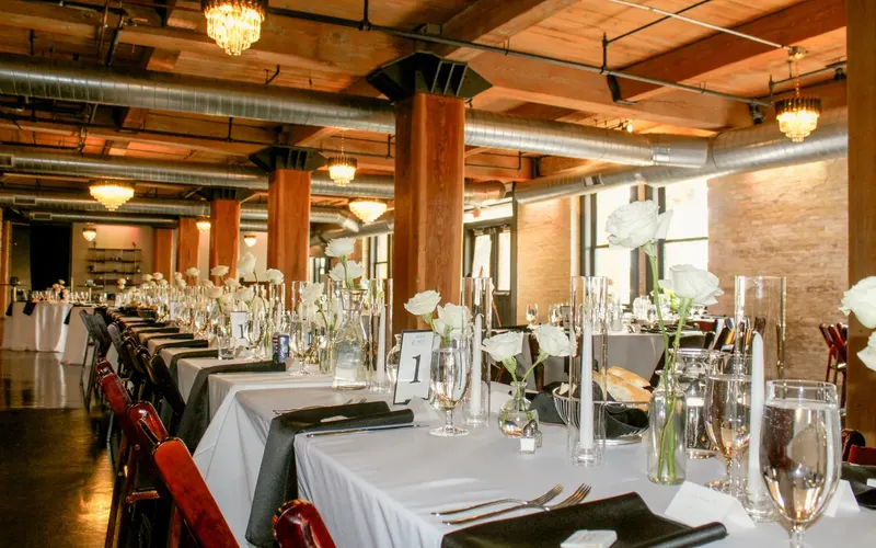 A long table set with white cloths and glassware in a brick and timber loft under chandeliers, before the guests arrive