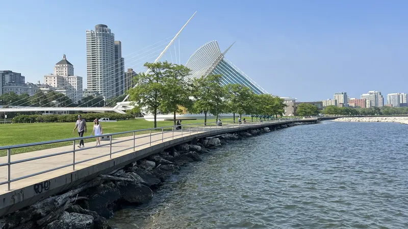 A paved lakefront path beside Lake Michigan with two walkers, trees, downtown towers and the white wings of the Art Museum beyond
