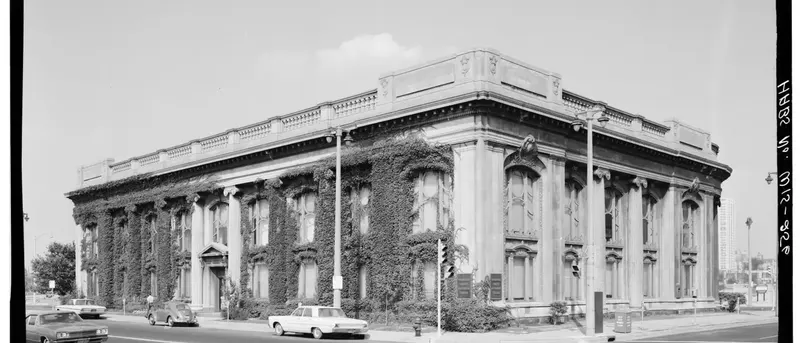 Black and white photograph of the limestone Second Ward Savings Bank building at the corner of two streets, partly covered in ivy