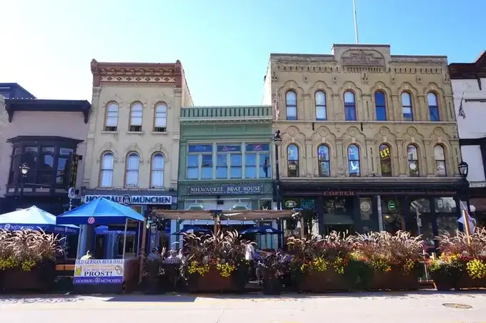 Three restored Victorian storefronts in cream, green and tan brick with arched upper windows, a sidewalk patio and planters in front