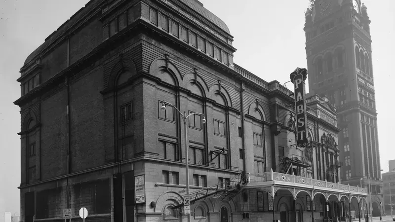 A black and white photograph of the Pabst Theater, a tall brick building with an arched entrance, a vertical PABST sign and a clock tower beside it
