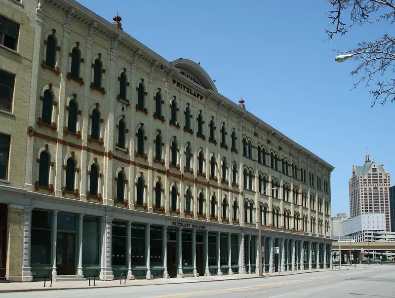 A long cream brick building with rows of arched windows and PRITZLAFF lettered on its center gable under the date 1875