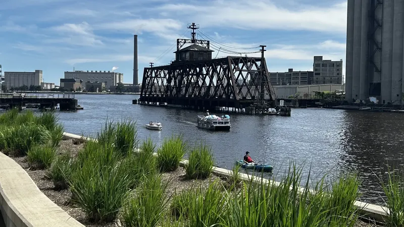 The Milwaukee River at Trestle Park, with tall grasses in the foreground, a black railroad swing bridge, small boats and a kayaker on the water