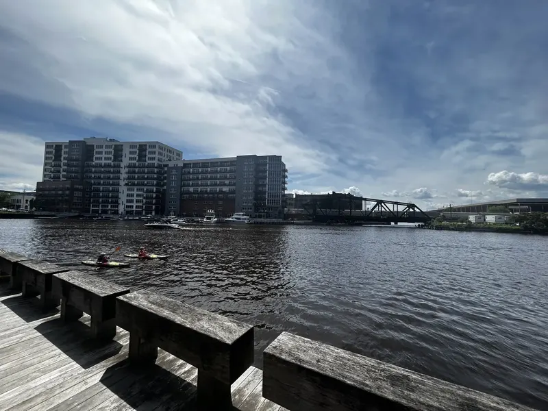 The Milwaukee River in the Third Ward, with a boardwalk railing, two kayakers on the water and apartment buildings on the far bank