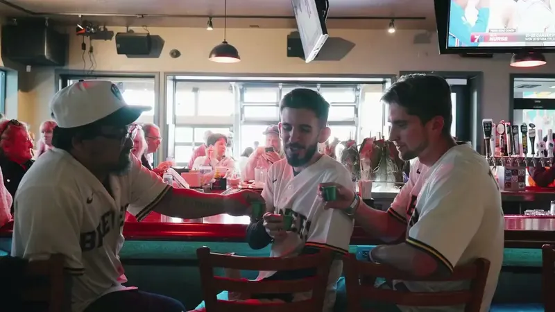 Three men in Brewers jerseys and caps clinking small glasses at a red bar with windows behind them