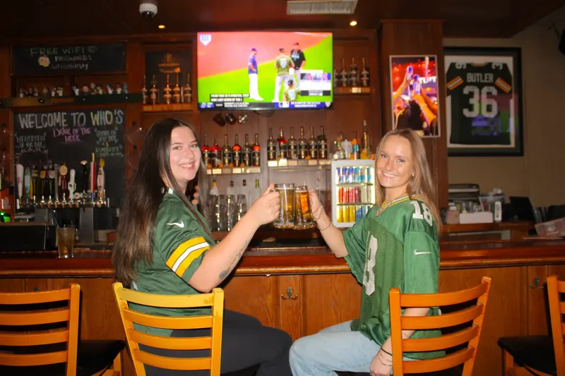 Two guests in Packers jerseys raising beers at the bar inside Who's on Third with the game on the screen