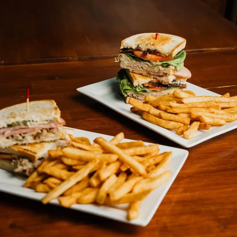 Two stacked deli sandwiches with fries on white plates on a dark wood table
