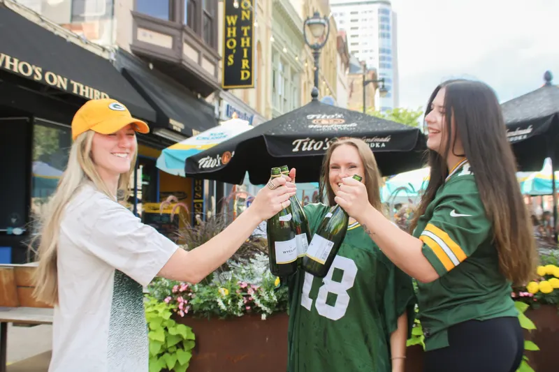 Three friends clinking bottles on the patio outside Who's on Third under the umbrellas